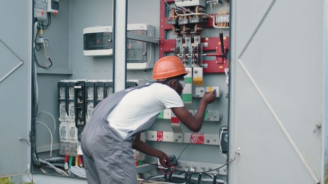african-american-man-in-overalls-safety-helmet-and-glasses-working-on-switchgear-at-so-SBI-351269919